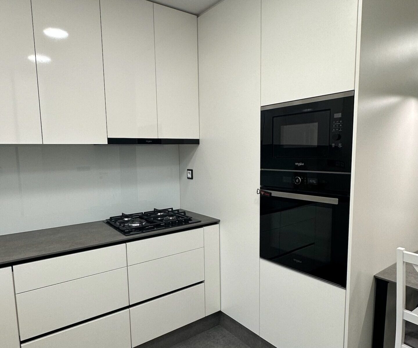 Moderní kuchyně s lesklými bílými skříňkami, tmavou pracovní deskou, vestavěným plynovým sporákem, nástěnnou troubou a mikrovlnnou troubou. V malém stolku vpravo je zastrčená bílá židle a podlaha je pokryta tmavě šedými dlaždicemi.